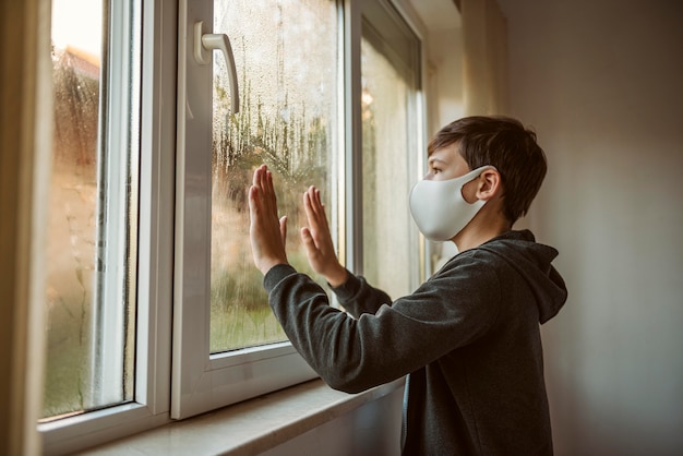 Free Photo side view little boy with face mask looking through the window