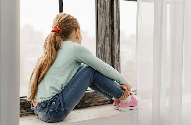 Free photo side view little girl sitting on a window sill