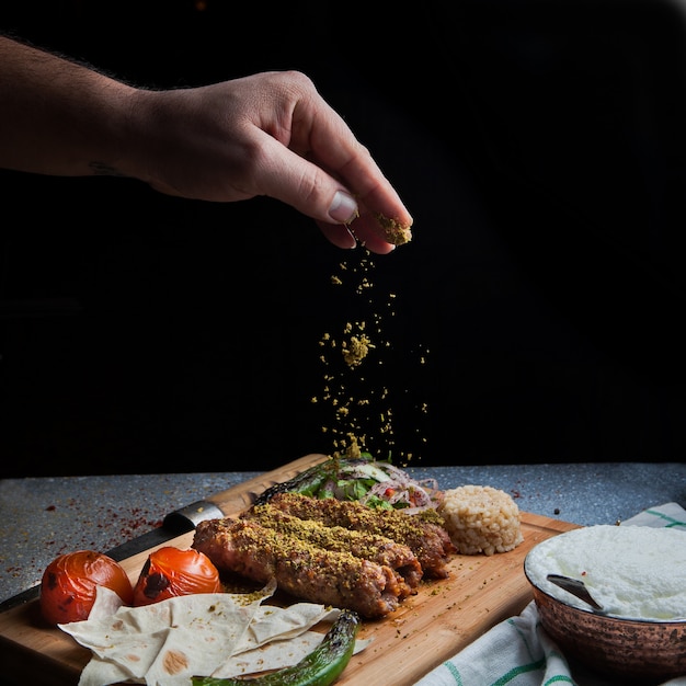 Free photo side view lule kebab with tomato and paper and ayran and hand adds spices in serving board