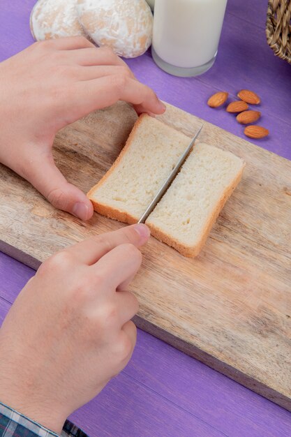 Side view of male hands cutting bread slice on cutting board with almonds gingerbreads milk on purple surface