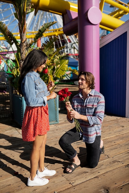 Side view of man getting down on his knee and offering bouquet of roses to girlfriend at the amusement park