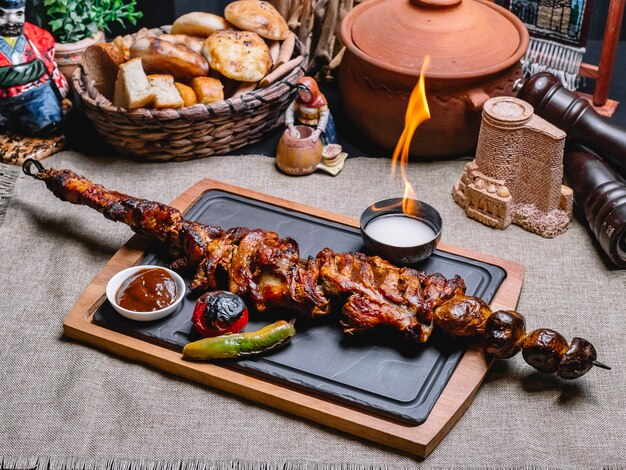 Side view meat kebab on a skewer with grilled potatoes and vegetables with sauce and fire on a board