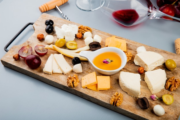 Free photo side view of melted butter with different kinds of cheese grape olive nuts on cutting board and lying glass of wine on white