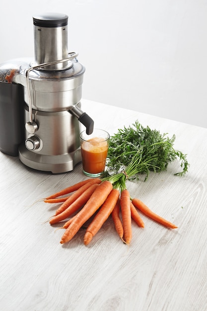 Free photo side view metallic professional juicer with glass filled with tasty juice for breakfast from organic farm carrots lying on wooden table.