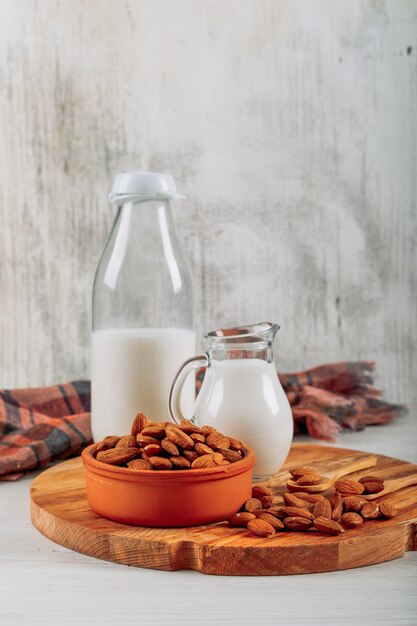 Side view milk carafe and bottle with bowl of almonds on wooden board on white wooden background. horizontal