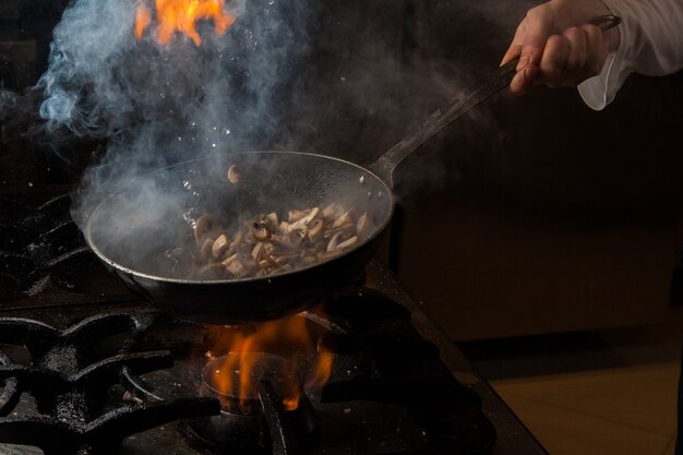 Side view mushroom frying with smoke and fire and human hand and pan in stove