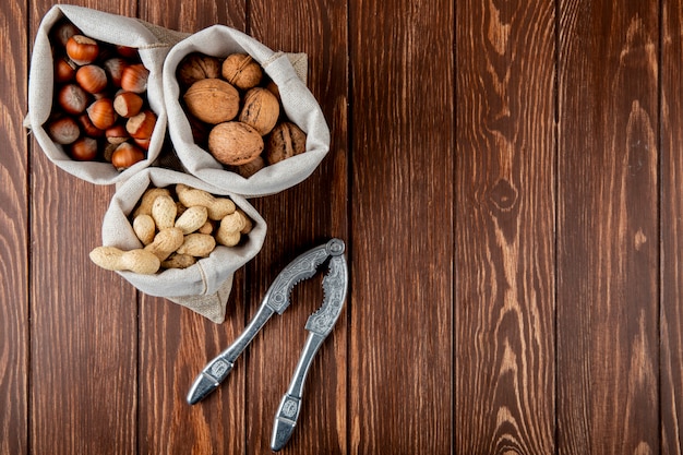 Side view of nuts in sacks walnuts peanuts and hazelnuts in shell with nut cracker on wooden background with copy space