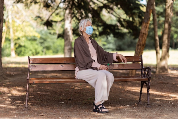 Side view of older woman with medical mask sitting on bench outdoors at nursing home