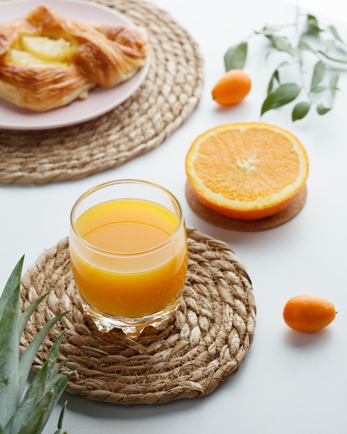 Side view of orange juice on trivet with kumquats cut orange croissant with leaves on white background