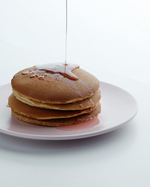 Free photo side view of pancakes in plate and strawberry jam syrup pouring on them on white background