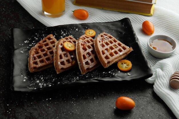 Side view of pancakes with kumquat slices in plate and jam orange juice on cloth on black background