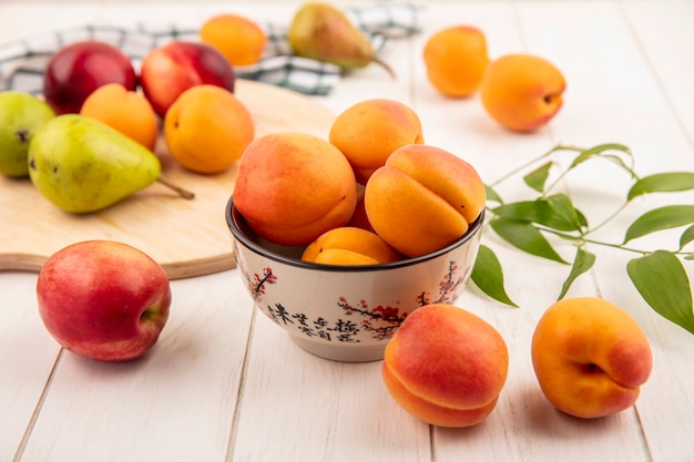 Side view of peaches in bowl and pears with peaches on cutting board and leaves on wooden background