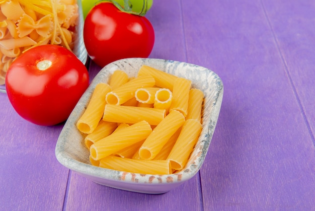 Side view of penne macaroni and other types in bowls and tomatoes on purple table with copy space