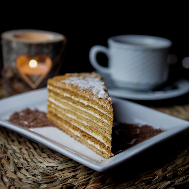 Free photo side view piece of honey cake with cup of tea and candle and white plate in serving napkins