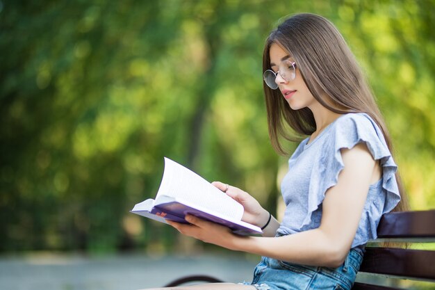 Side view of pleased brunette woman in eyeglasses sitting on bench and reading book in park