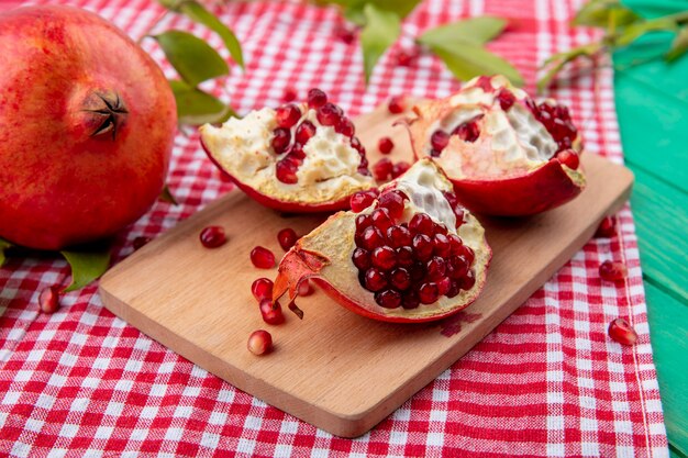 Side view of pomegranate pieces on cutting board with whole one and leaves on plaid cloth and green