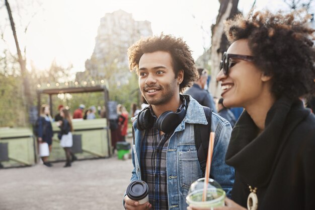 Side-view portrait of charming african-american boyfriend with afro haircut looking aside while walking with his girlfriend in park, drinking coffee and enjoying warm evening