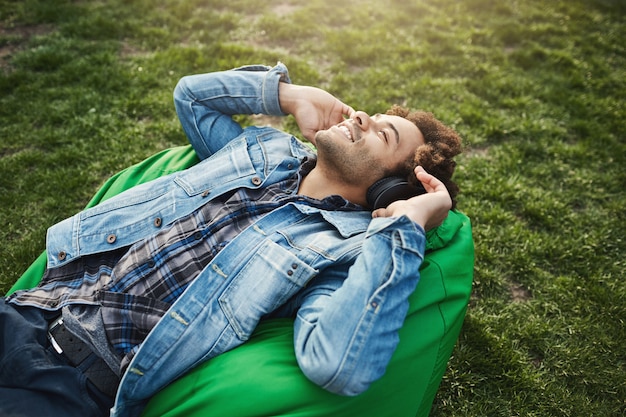 Free Photo side-view portrait of happy attractive african-american with afro hairstyle lying on bean bag chair while listening music and looking at sky with relaxed and pleased expression, spending time in park