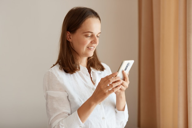 Side view portrait of positive woman with dark hair wearing white cotton shirt standing with cell phone in hands near window in living room at home, browsing internet.