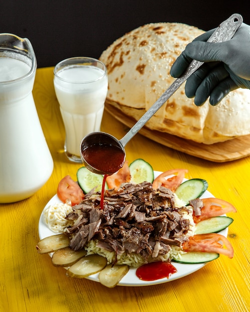 Free Photo side view of pouring sauce on doner on plate served with pickles fresh cucumbers and tomatoes