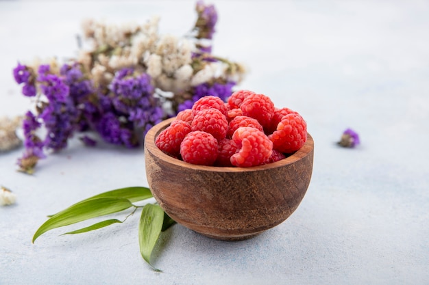 Side view of raspberries in bowl with flowers and leaves on white