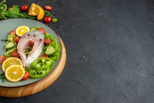 Side view of raw fish and fresh chopped vegetables lemon slices spices on a gray plate on a round board on black distressed surface