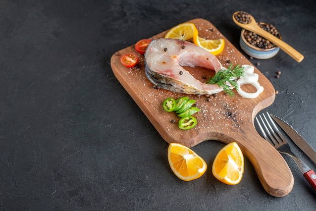 Free photo side view of raw fish and fresh chopped vegetables lemon slices spices on a wooden board cutlery set on black distressed surface