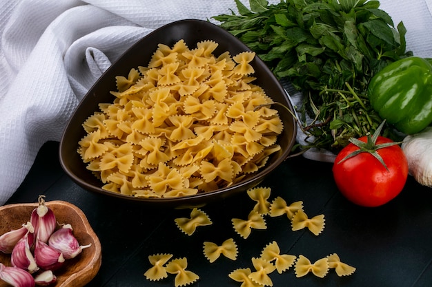 Side view of raw pasta in a bowl with tomatoes garlic and bell pepper with mint on a white towel on a black surface