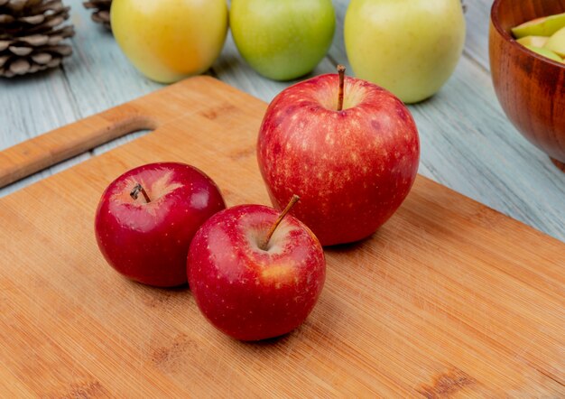 Side view of red apples on cutting board with yellow and green ones on wooden background