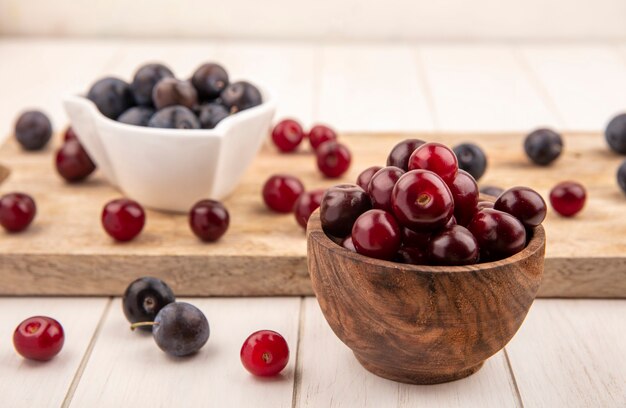 Side view of red cherries on a wooden bowl with dark purple sloes on a white bowl on a wooden kitchen board on a white wooden background