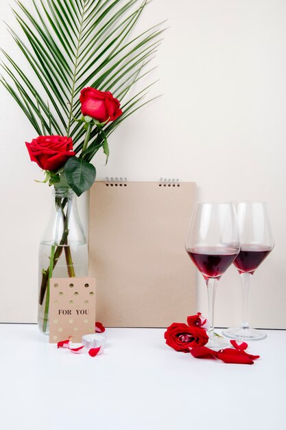 Side view of red roses with palm leaf in a glass bottle standing near a sketchbook and two glasses of red wine on white background