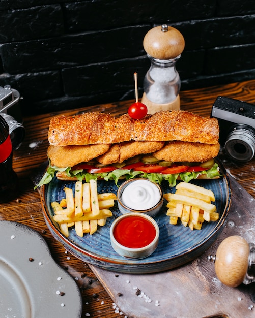 Side view of sandwich with chicken nuggets salad leaves pickles and sauce served with french fries on wooden table