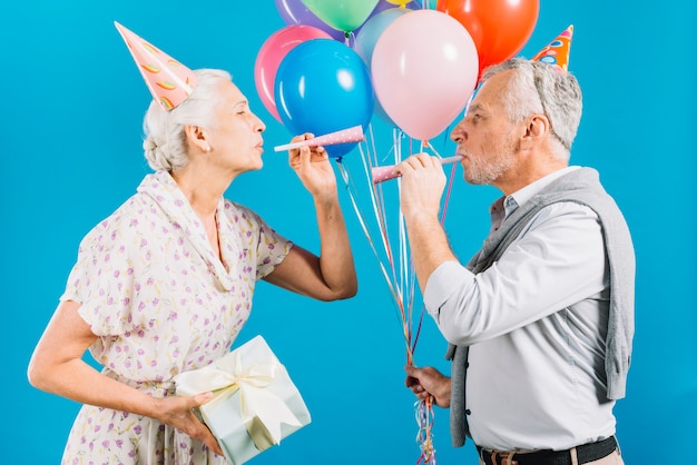 Side view of senior couple looking at each other blowing party horn on blue backdrop