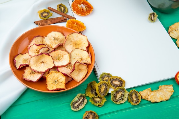 Side view of a sketchbook with dried apple chips on a plate on white and green background