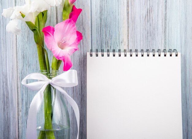 Side view of a sketchbook with white and pink color gladiolus flowers in a glass bottle on grey wooden background