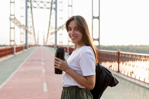 Side view of smiley traveling woman holding thermos on bridge