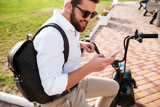 Side view of Smiling Bearded man in sunglasses sitting on modern motorbike outdoors and using smartphone