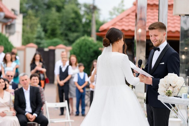 Side view smiling groom man with microphone holding bride's hand swears an oath on wedding ceremony outdoors Happy guests on background enjoying beautiful couple Elegant wife in puffy dress