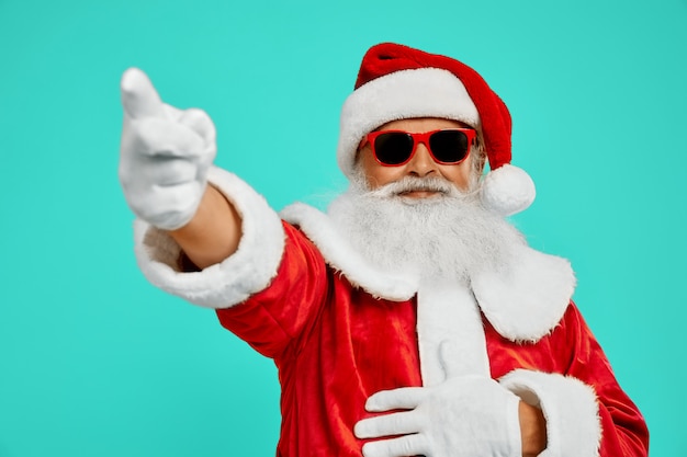 Free photo side view of smiling man in red santa claus costume. isolated portrait of senior male with long white beard in sunglasses pointing away