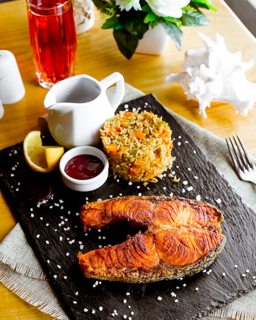 Free photo side view steak from fried red fish with rice with vegetables a slice of lemon and pomegranate sauce on the board