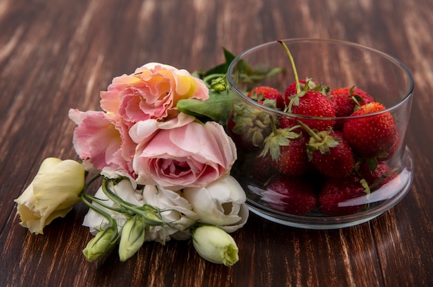 Free photo side view of strawberries in bowl and flowers on wooden background