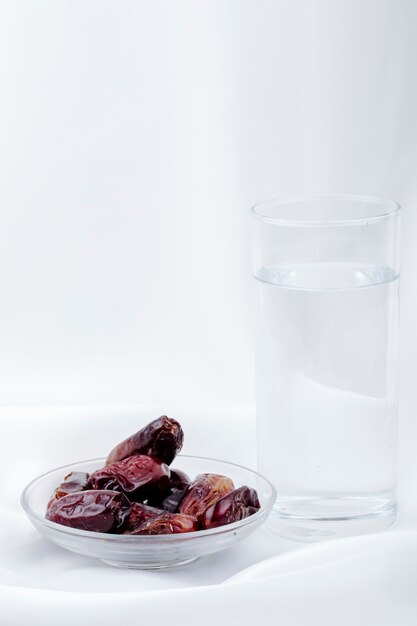 Side view of sweet dried date fruits in a saucer with a glass of water at white background