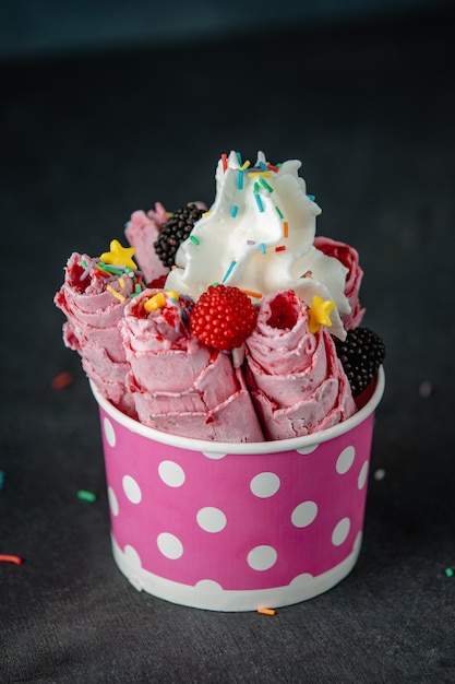 Side view of thai ice cream roll under whipped cream decorated with berries and colorful sprinkles in a cardboard bowl on black wall