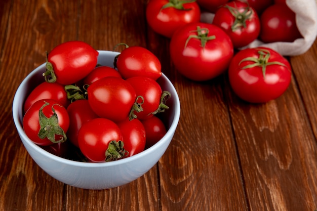 Side view of tomatoes in bowl with other ones spilling out of sack on wooden table