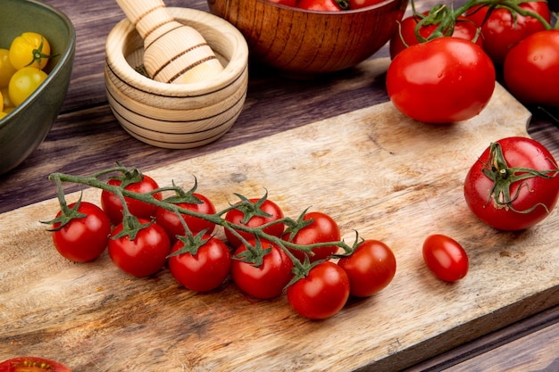 Side view of tomatoes on cutting board with other ones garlic crusher on wooden table