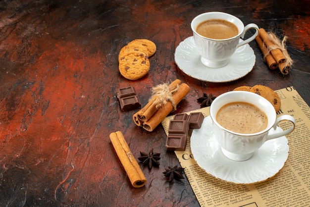 Side view of two cups of coffee cookies cinnamon limes chocolate bars on an old newspaper on the left side on dark surface