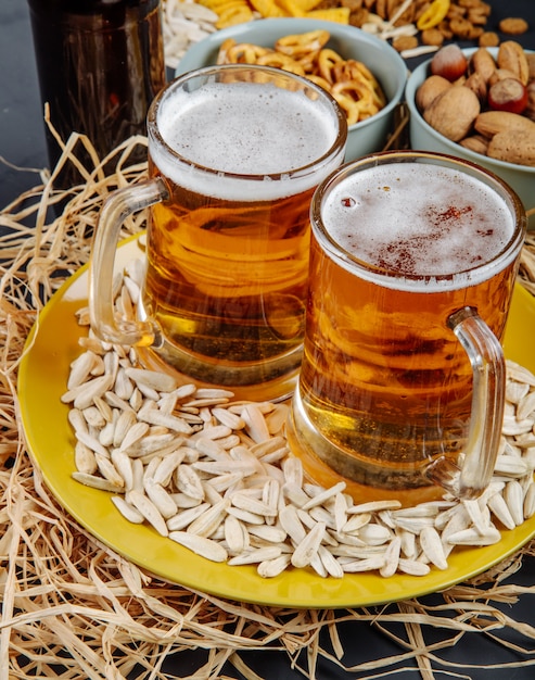 Free photo side view of two mugs of beer on a plate with sunflower seeds on straw