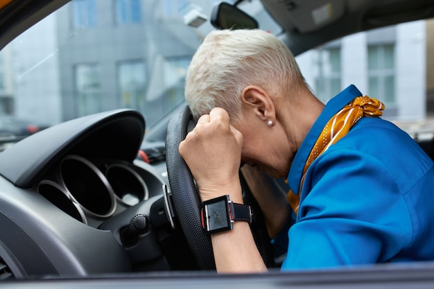 Free photo side view of unhappy stressed middle aged woman squeezing fists and resting head on steering wheel, stuck in traffic jam, being late for work or get into car accident, sitting in driver's seat