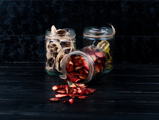 Side view of various dried fruit slices in glass jars strawberry banana and kiwi on black background