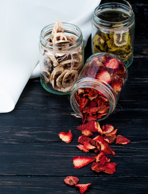 Side view of various dried fruit slices in glass jars strawberry banana and kiwi on black background